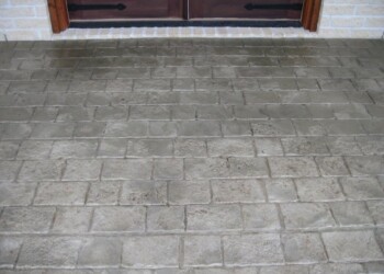Concrete porch with stamped overlays and brick border in front of a wooden door. Stamped concrete porch with brick border and wooden door in the background.