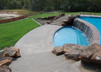 Beautiful stamped concrete pool deck with rock accents and waterfall in a backyard. Stamped concrete pool deck with rock accents and waterfall in a backyard.
