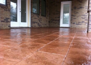Finished stamped concrete patio in a reddish-brown color outside a residential home. Stamped concrete patio with a reddish-brown finish outside a brick house.