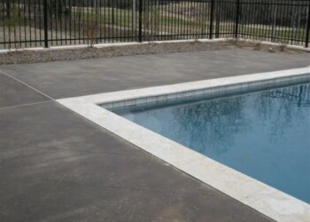 Concrete pool deck with acid staining near a swimming pool in a backyard. Concrete pool deck with acid stained finish and black metal fence in the background.
