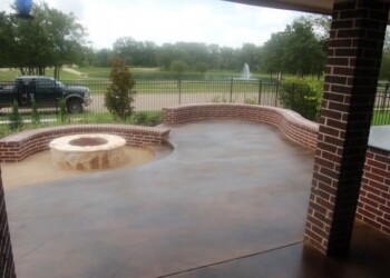 Finished concrete patio with brick wall and fire pit in a residential backyard. Concrete patio with brick wall and fire pit, overlooking a park with trees and a fountain.