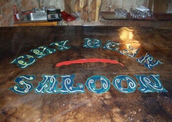 Metal letters spelling 'WELCOME SALOON' arranged on a wooden bar in a rustic setting. Custom engraved metal letters spelling 'WELCOME SALOON' on a wooden bar surface.