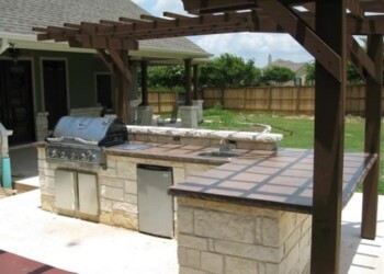 Outdoor kitchen with stone countertops and built-in grill in Euless, Texas. Outdoor kitchen with stone countertops and built-in grill under a wooden pergola in Euless, Texas.