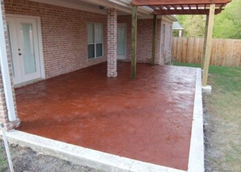 Stamped overlays on a concrete patio at a home in Euless, Texas. Stamped concrete patio with a red-brown finish under a covered porch in a residential backyard.