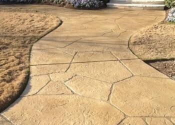 Stamped overlays on concrete walkway in front of a house with steps and a wooden door. Stamped concrete walkway leading to a house entrance with bushes and flowers.
