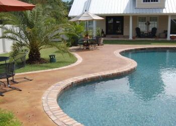 A backyard with a kidney-shaped pool, patio furniture, and a white house with a metal roof. Backyard with a kidney-shaped pool, patio, and a white house with a metal roof in a sunny setting.