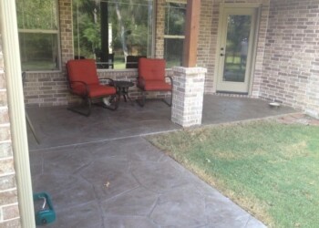 Stamped overlays on concrete patio with seating area in a backyard. Stamped concrete patio with red cushioned chairs and brick columns in a residential backyard.