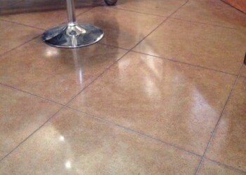 Polished concrete floor in a room with a metal table and wooden baseboard. Polished concrete floor with visible grout lines in a room corner.
