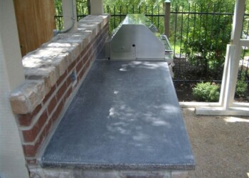 Outdoor concrete countertop with a built-in grill in a backyard setting. Concrete countertop installed outside next to a brick wall and black metal fence.