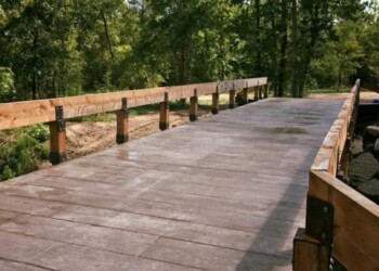 Concrete bridge with wooden railings in a wooded area, part of a construction project. Concrete bridge under construction with wooden railings and green trees in the background.