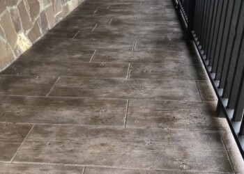 Stamped overlays on porch with wood texture and black railing in a residential area. Stamped concrete porch with wood-like finish and black railing in a residential setting.