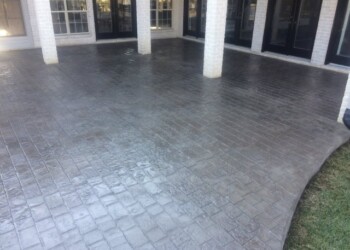 Stamped overlays on concrete patio at a residential property in Eads, Tennessee. Stamped concrete patio with a dark gray finish and brick pattern in front of a modern house.