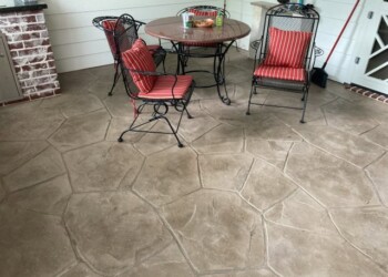 Stamped overlays on concrete patio with outdoor table and chairs in a backyard. Stamped concrete patio with outdoor furniture and red-striped cushions in a backyard.