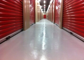 Storage units with red roll-up doors in a clean, well-lit facility. Long hallway with red storage unit doors on both sides in a storage facility.
