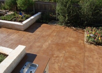 Stamped overlays on concrete patio in a backyard with garden beds and greenery. Stamped concrete patio with garden beds and plants in a backyard.