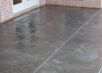 Stamped overlays on a concrete patio at a residential property in Euless, Texas. Stamped concrete patio with a smooth, dark finish near a brick house.