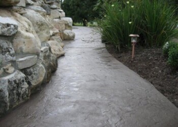 Stamped concrete walkway beside a stone wall and garden in a residential yard. Concrete walkway with stamped overlay next to a stone wall and garden in a residential yard.